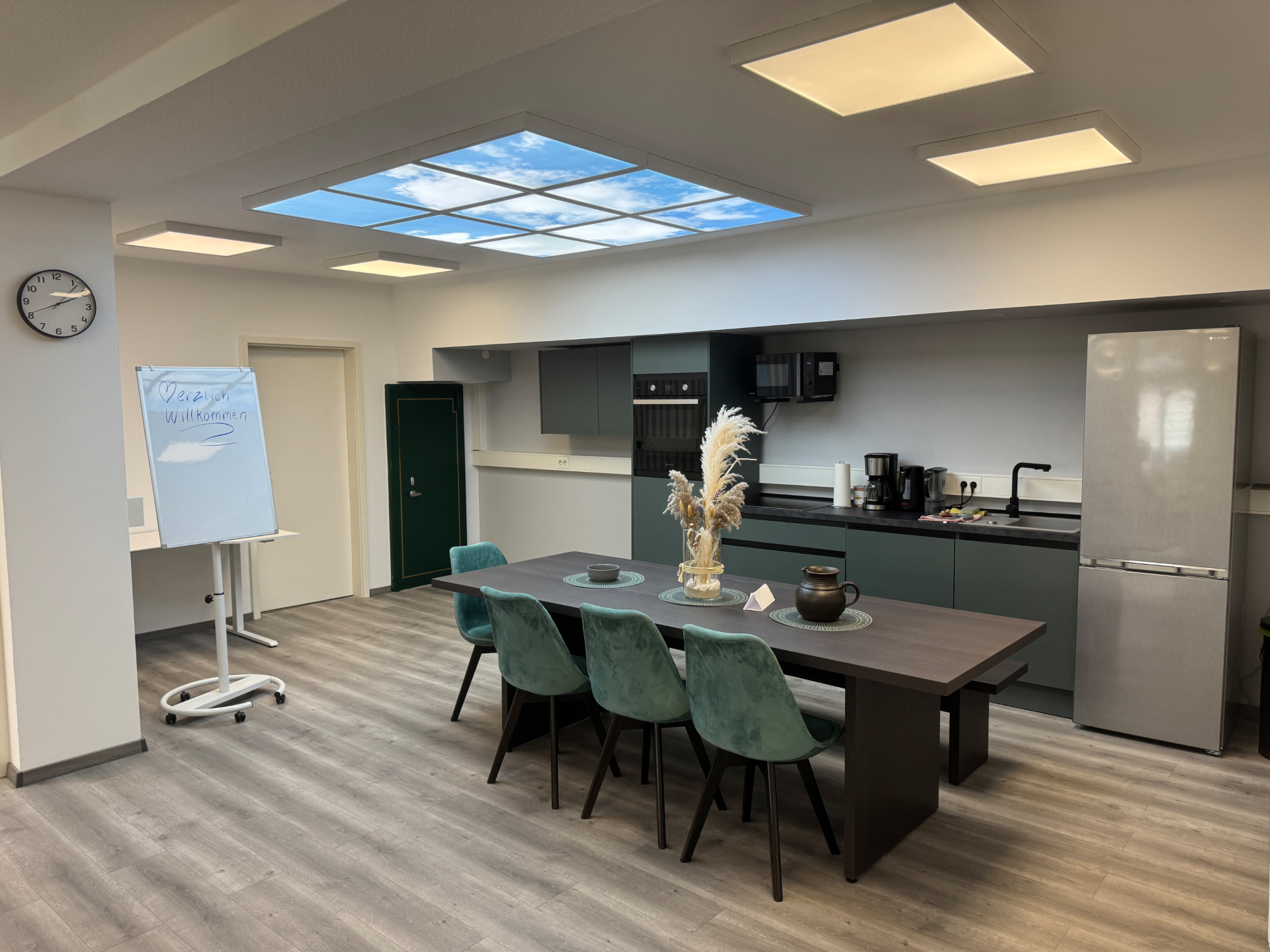 Gemeinschaftsraum und Küche im Coworking Space CoWork Station Bakede: großer Esstisch mit türkisen Stühlen und Sitzbank, dunkle Küchenzeile mit Backofen und Kühlschrank, Himmeldecken-Panel mit Wolkenmotiv