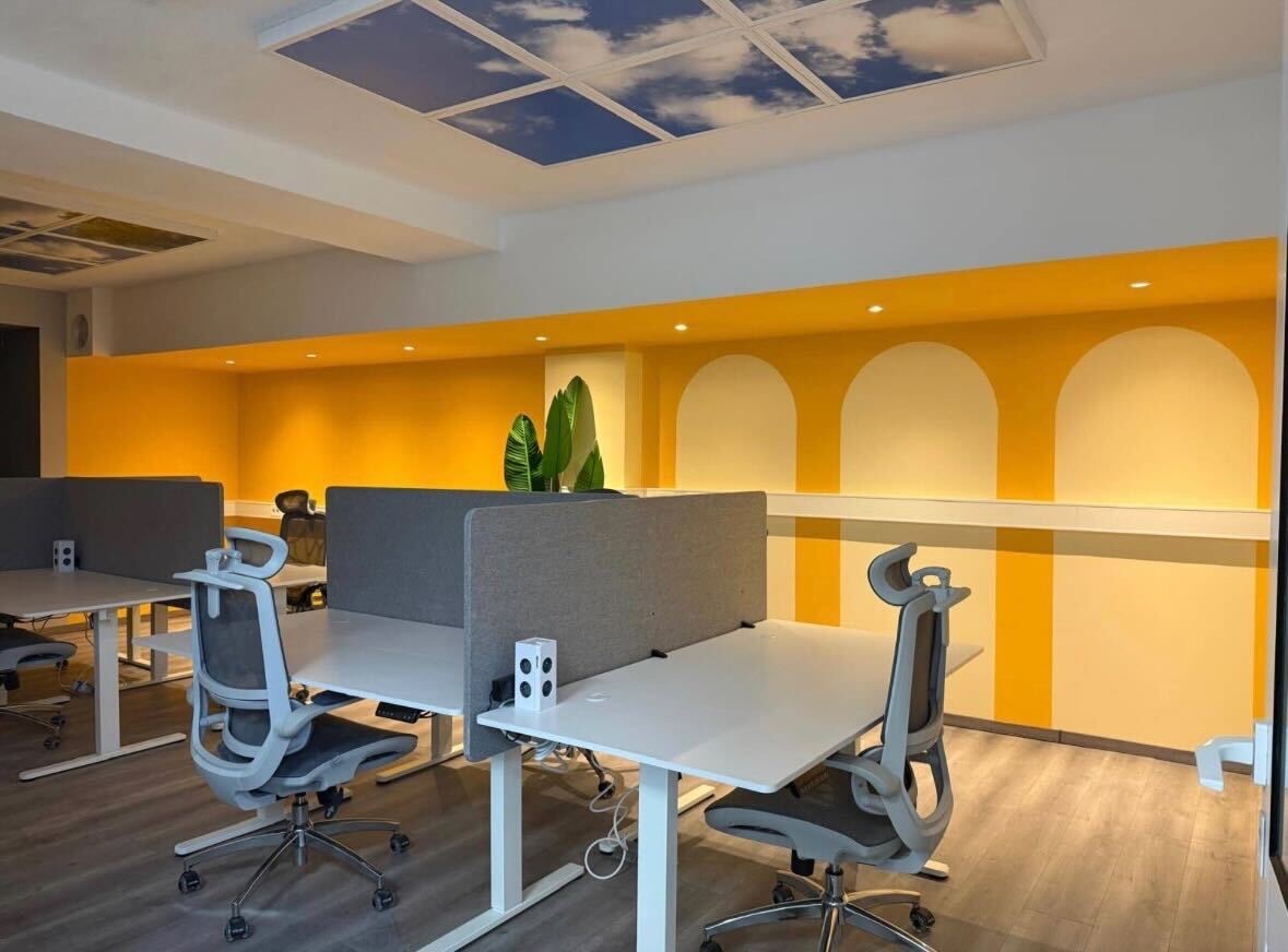 Open Space im Coworking Space CoWork Station Bakede: Arbeitsplätze mit höhenverstellbaren Tischen vor einer gelben Wand mit Rundbogennischen, Pflanzen und Himmeldecken-Panel